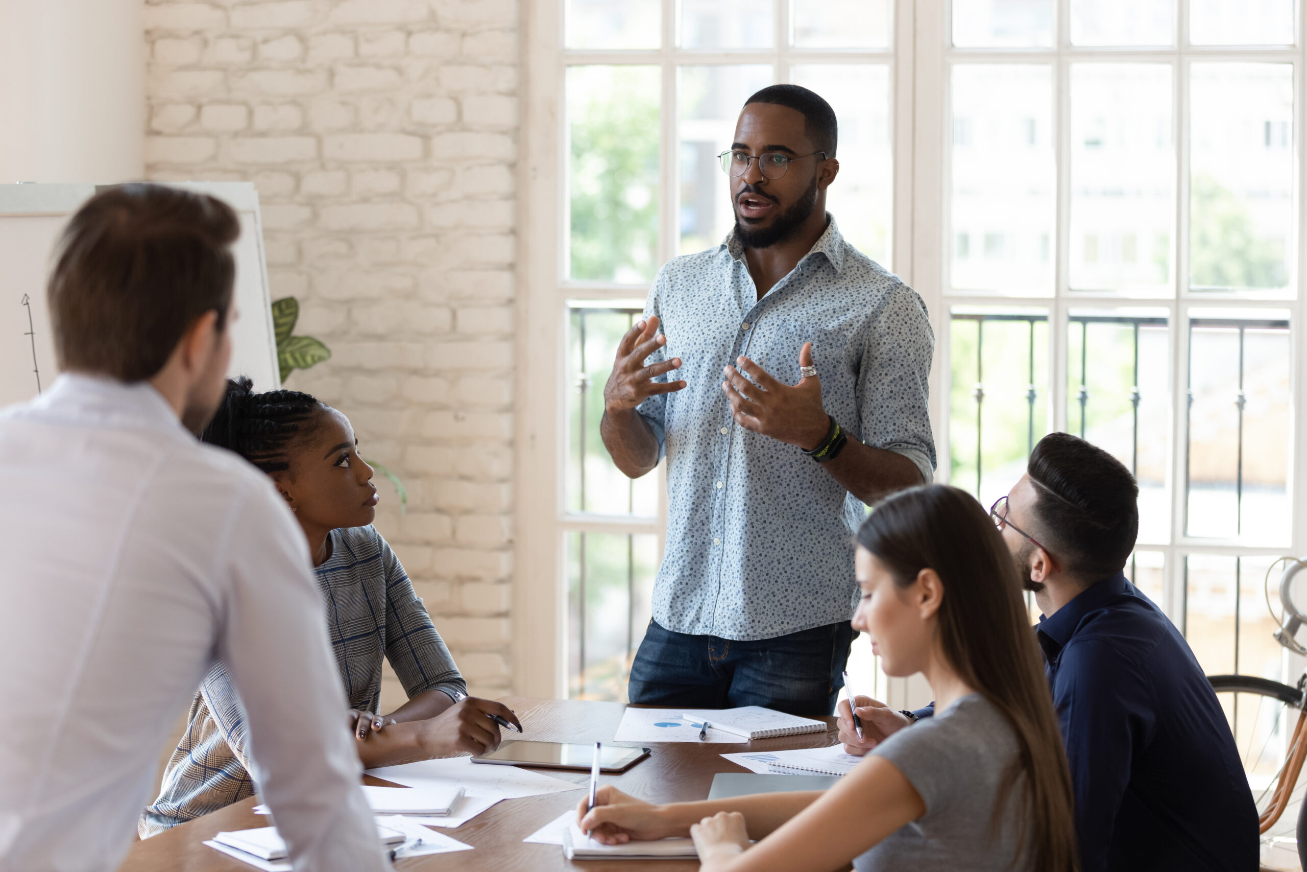 A people manager stands in an office at the head of a meeting with employees to discuss mental health benefits in the workplace.